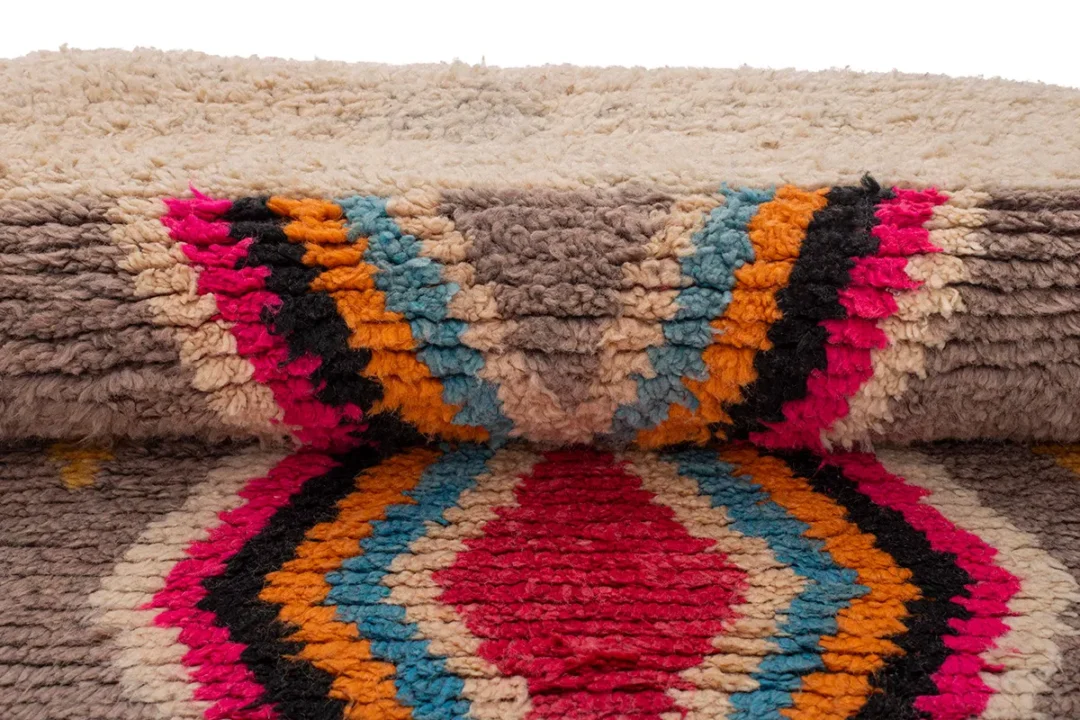 Close-up of a colorful geometric-patterned rug with textured piles. Features diamond shapes in red, blue, orange, pink, and black against a gray and beige background.