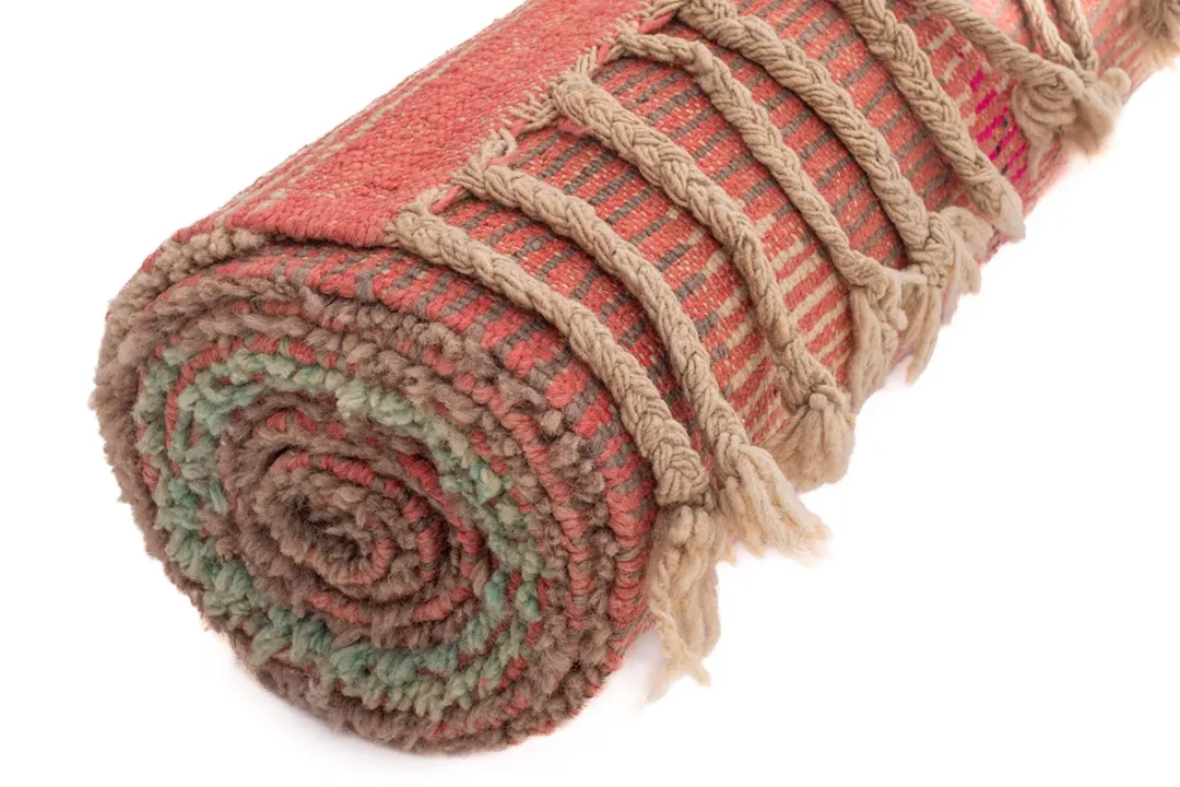 Rolled up woven rug with red, brown, and green hues, featuring fringed edges.