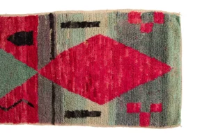 A rectangular rug with geometric patterns featuring red, black, and green colors, including a large central diamond shape on the left and smaller shapes on the right.