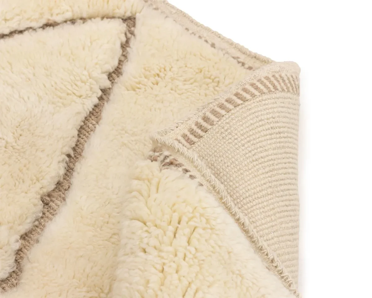 Close-up of a cream-colored, textured rug corner. The rug has a shaggy, soft pile with a contrasting woven backing and subtle geometric pattern.