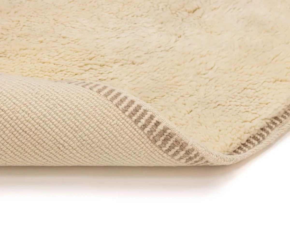 Close-up of a beige, textured rug with a fluffy upper surface and a woven underside, folded at the corner to display both textures.