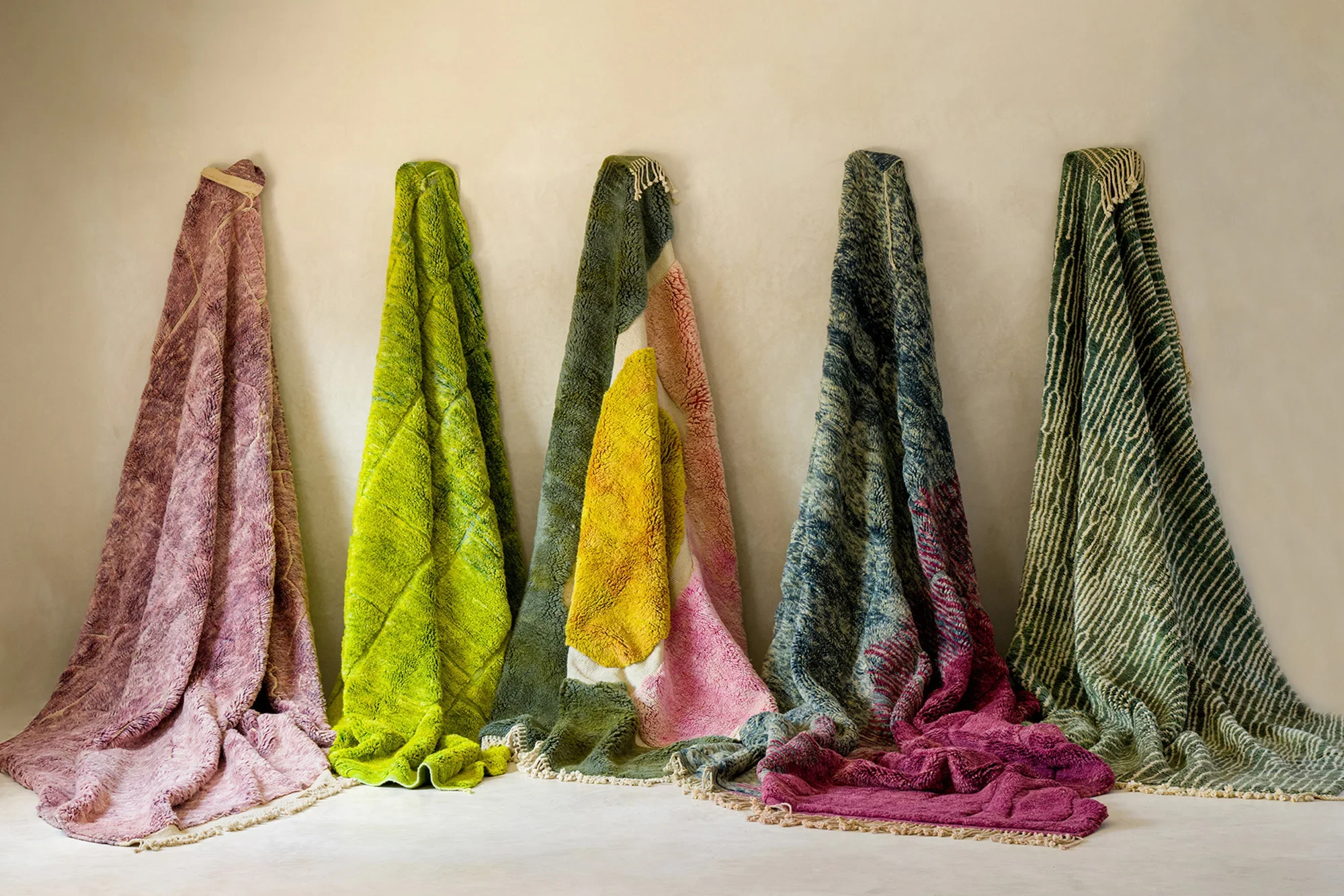 Five colorful textured rugs leaning against a wall, each with a unique pattern and color, including shades of pink, green, yellow, and blue.