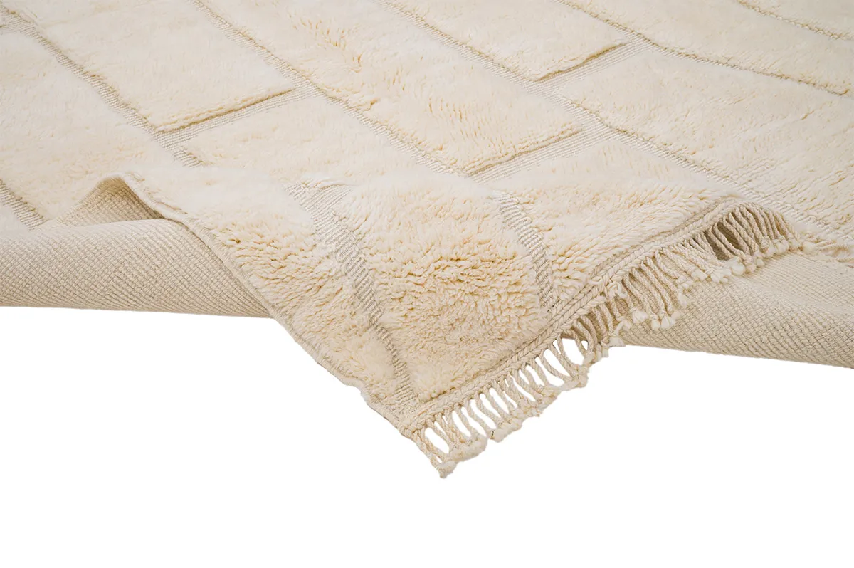 Close-up of a beige textured rug with fringe on the edges, lying on a plain surface.