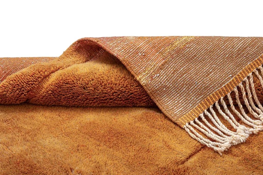 A folded orange and brown textured rug with a woven pattern and white fringed edge on a plain background.