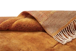 A folded orange and brown textured rug with a woven pattern and white fringed edge on a plain background.