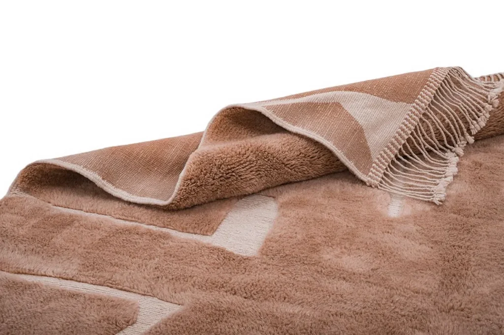A close-up of a folded brown and beige rug with a geometric pattern and fringed edge, displayed on a white background.