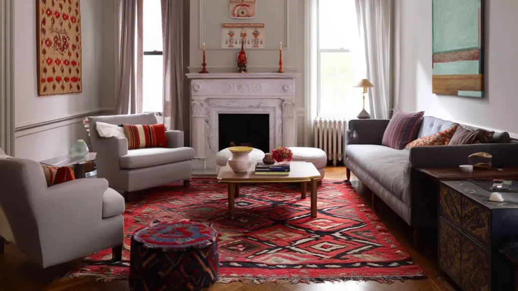 A living room with a marble fireplace, two armchairs, a sofa, and a colorful patterned rug. Decor includes cushions, vases, a lamp, and wall art. Bright daylight filters through the windows.