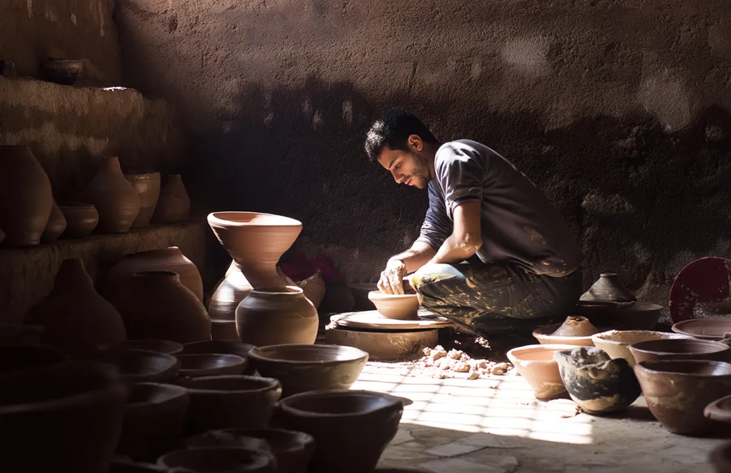A person is focused on shaping clay on a pottery wheel in a dimly lit workshop filled with various pottery pieces.