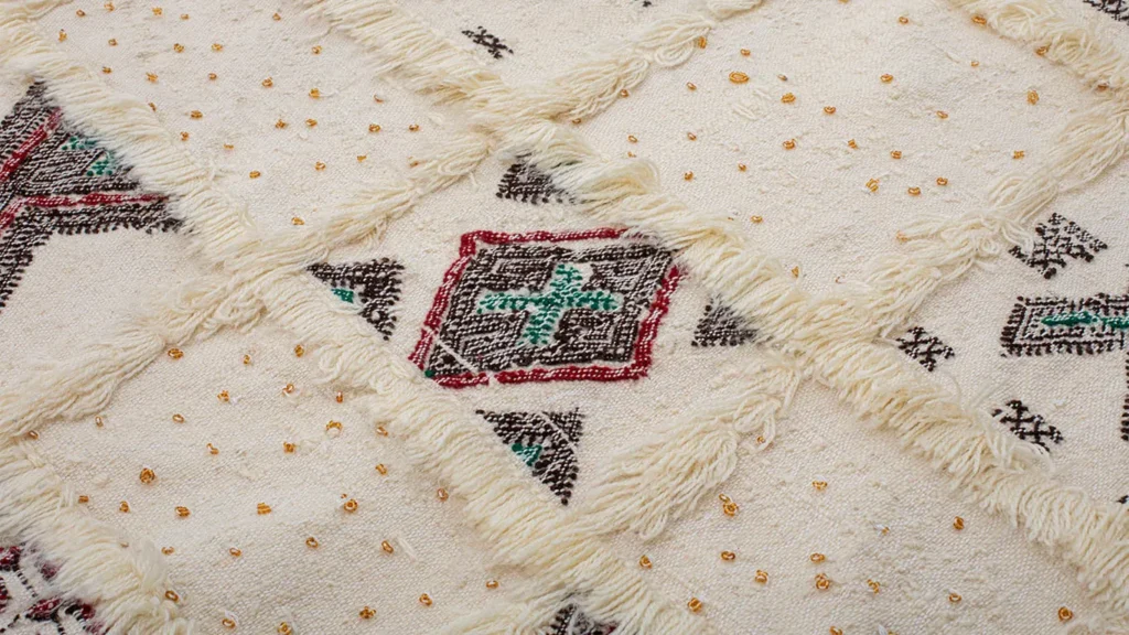 A textured cream rug with geometric patterns in red, teal, and black, adorned with small golden beads and raised fringes in a grid-like design.
