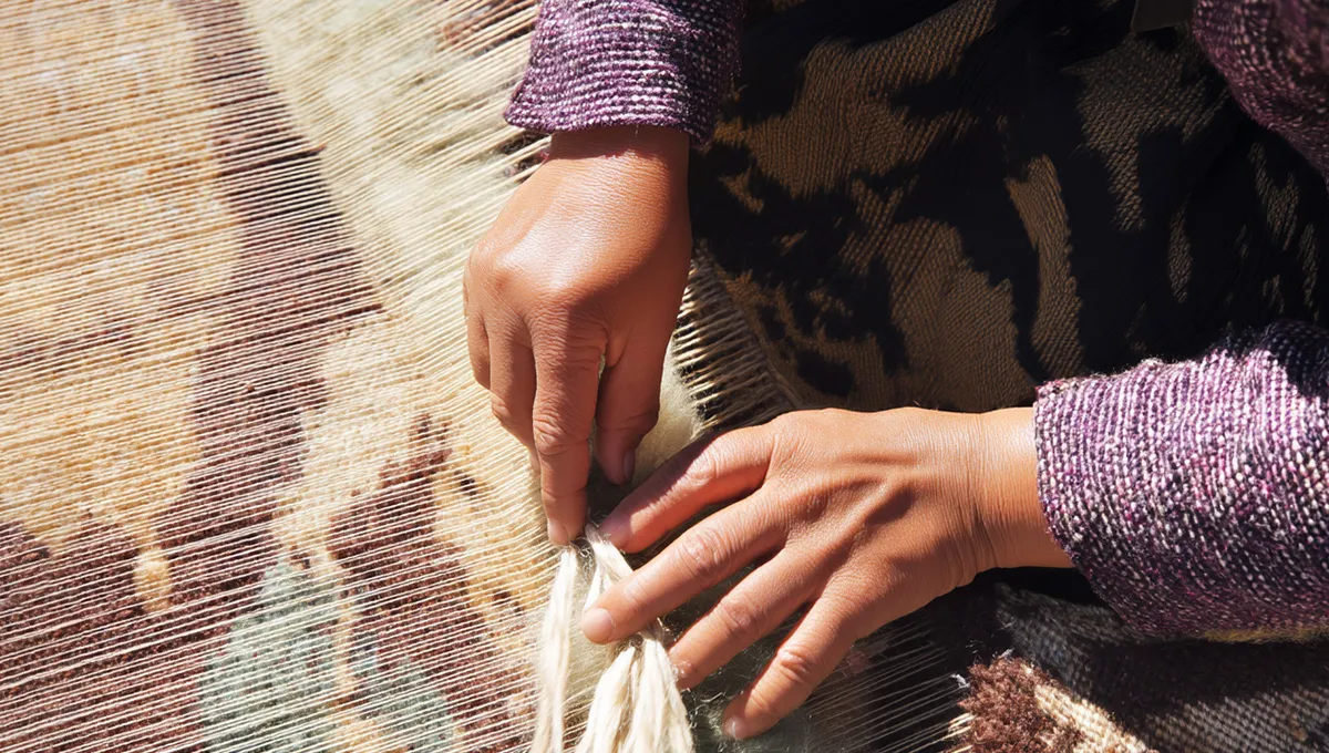 Close-up of hands weaving a rug on a loom. The person is tying knots using cream-colored thread against a backdrop of woven patterns. Theyre wearing a purple, textured long-sleeve.