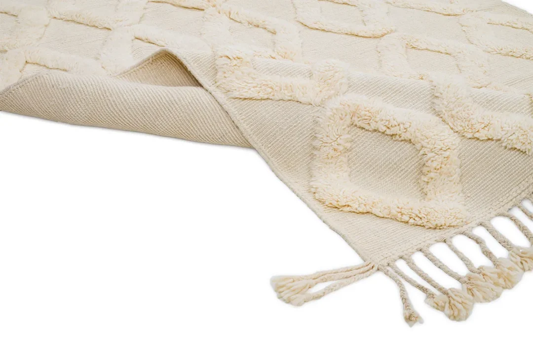 A cream-colored textured rug with a geometric pattern, featuring tasseled fringes on one edge, is laid out partly folded on a white background.
