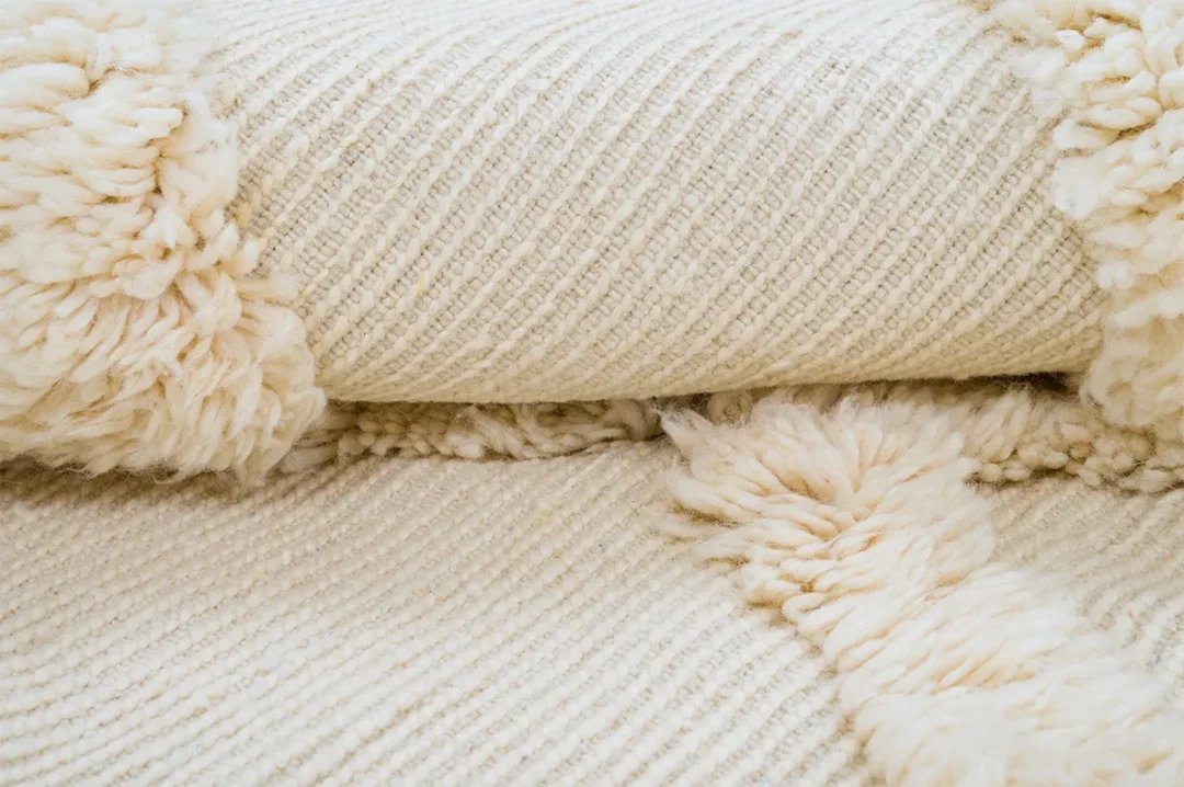 A close-up view of a folded, textured cream-colored wool rug with soft, fluffy sections.