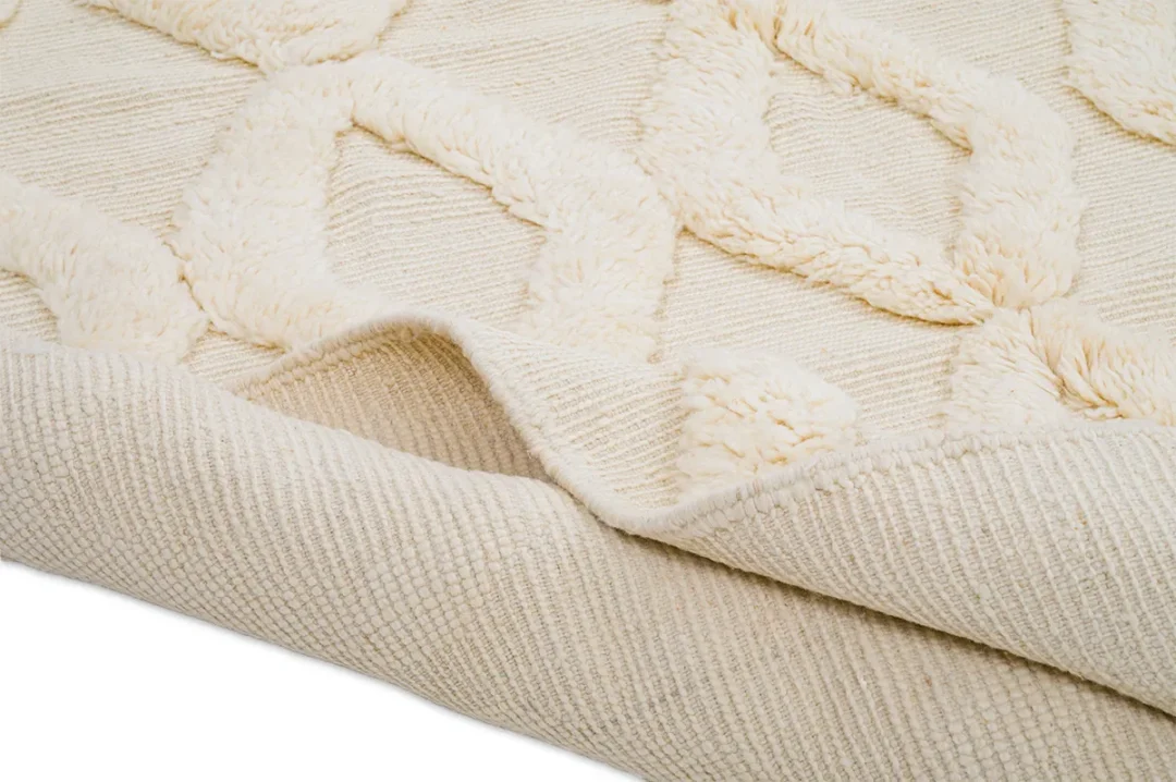 A close-up view of a cream-colored textured rug with a raised diamond pattern and one edge slightly folded over.
