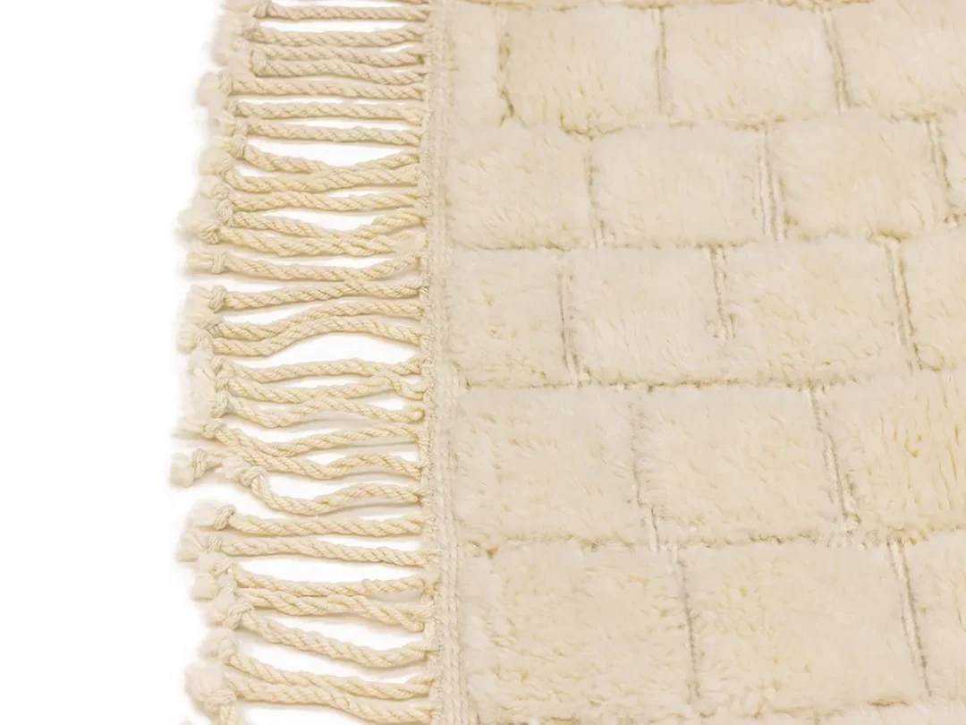 Close-up of a cream-colored, woven rug with a textured, square pattern and twisted fringe on one edge.