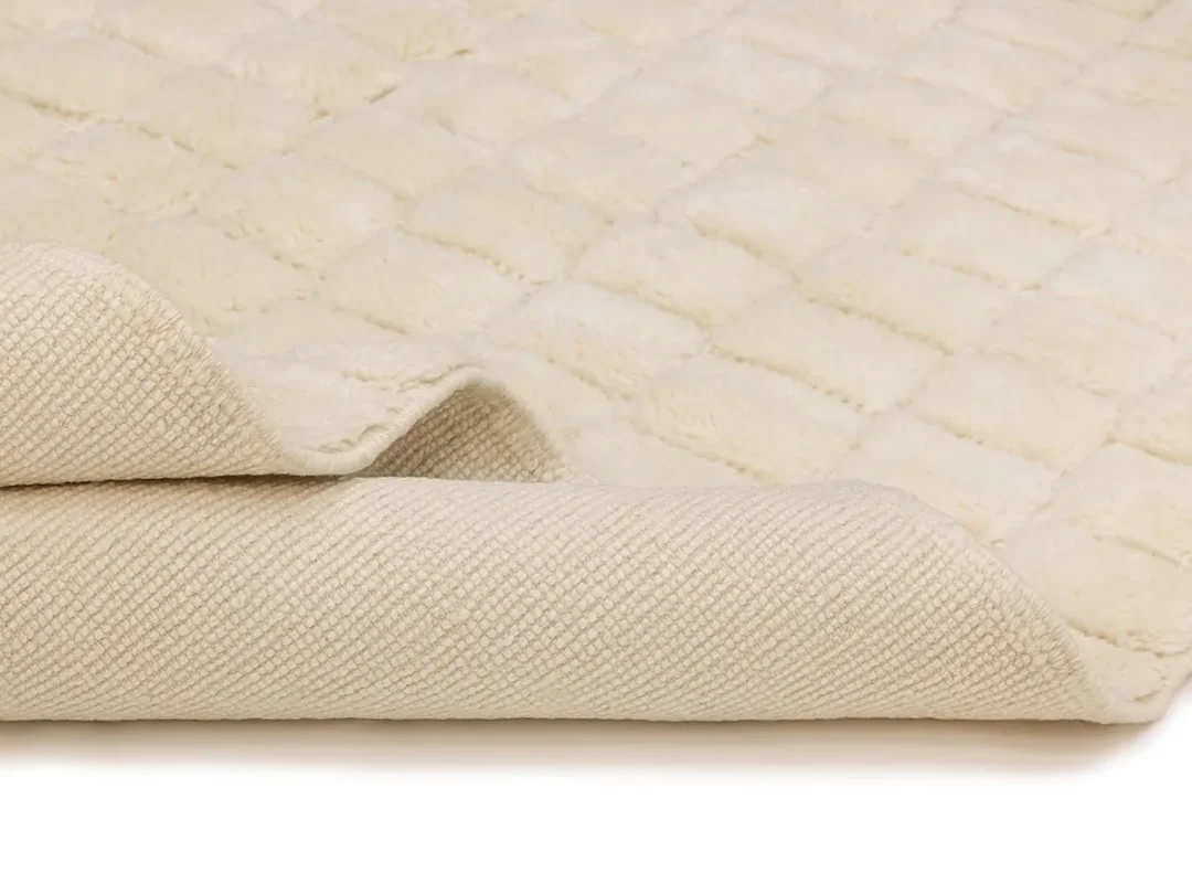 A close-up of a cream-colored textured rug with the corner partially folded over, showing both the soft upper surface and the woven underside.