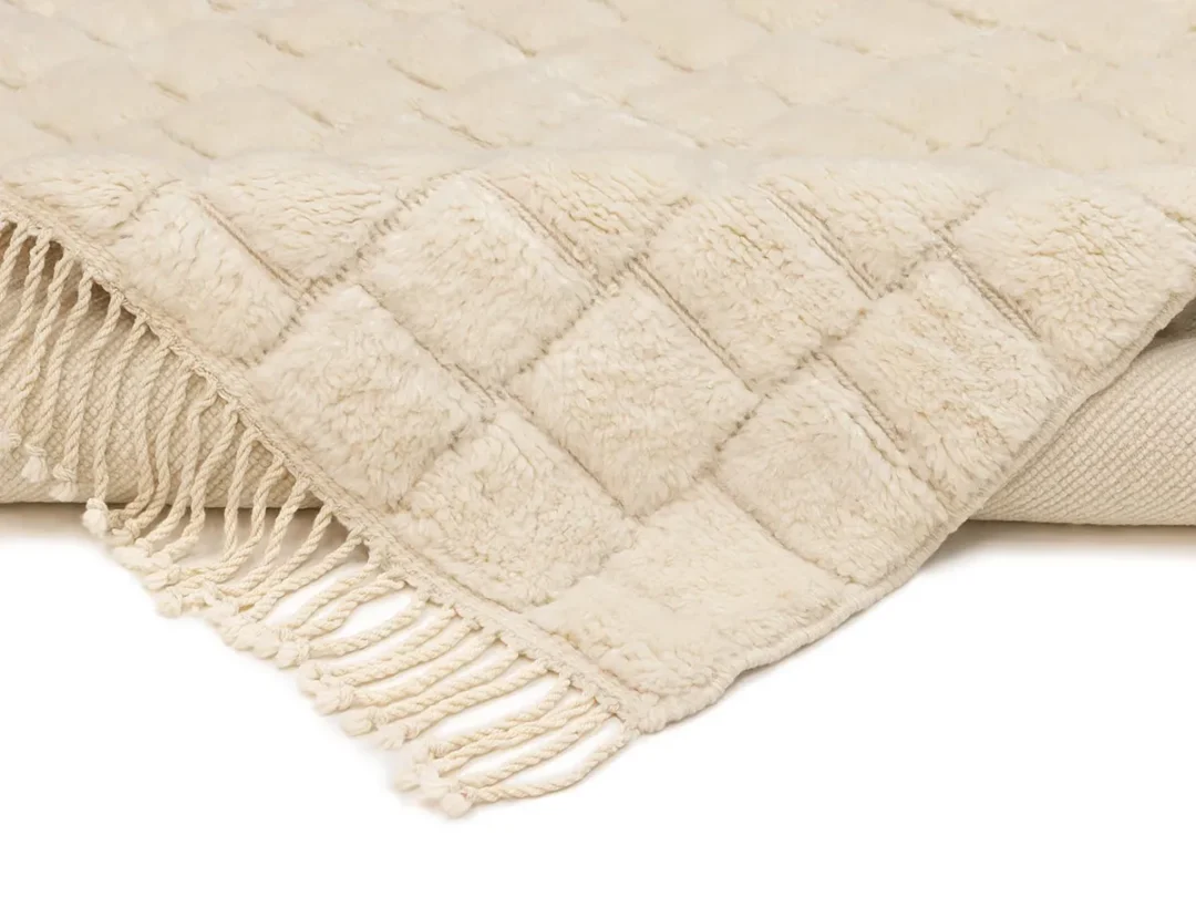 A cream-colored, textured throw blanket with a quilted pattern and fringed edges, folded over at the corner.