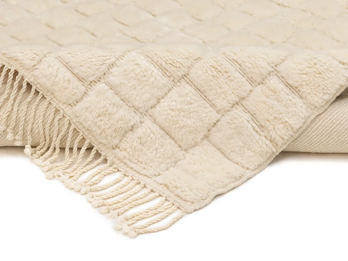 A cream-colored, textured throw blanket with a quilted pattern and fringed edges, folded over at the corner.