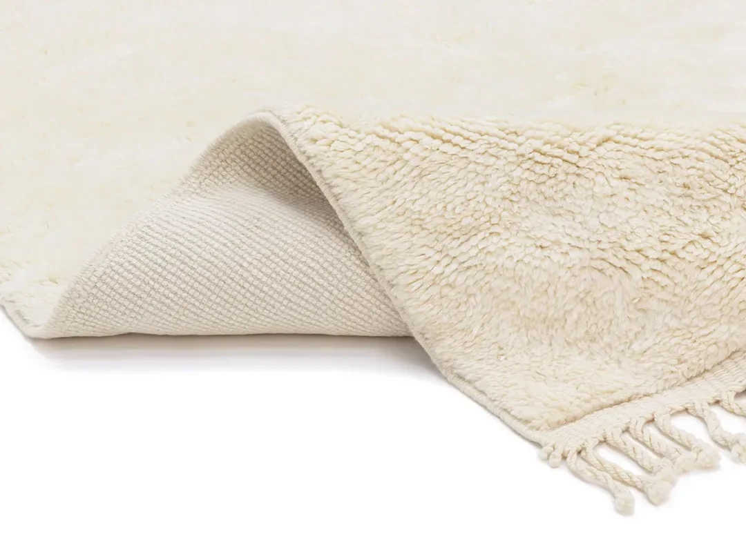 A cream-colored, textured rug is shown with one corner flipped over, revealing a woven underside and tassels on the edge.