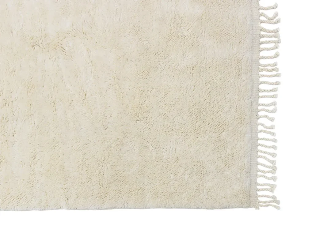 A close-up of a beige, textured rug with fringe detail along the right edge.