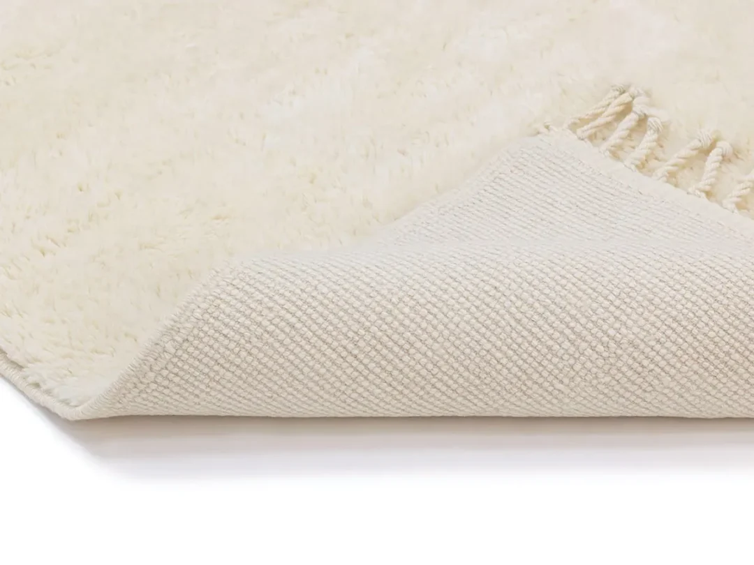 A close-up of a cream-colored textured rug with one corner folded to show the woven underside and fringed edge.