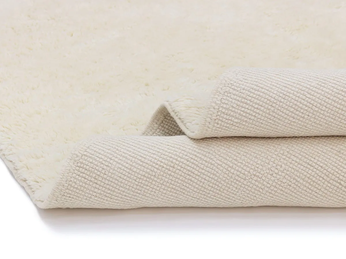 A close-up of a cream-colored textured rug with one edge folded over, showing the woven underside and soft pile surface.