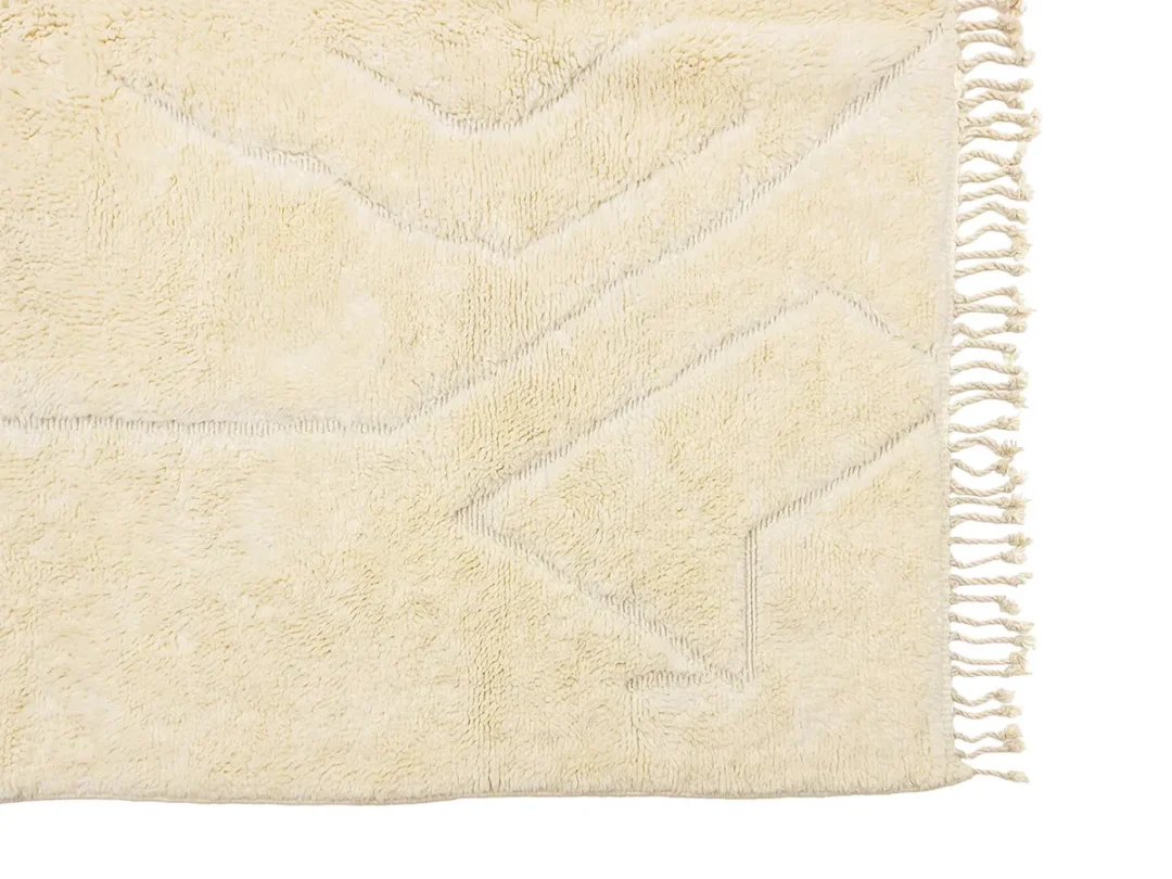Close-up of a beige textured rug with subtle geometric patterns and fringed tassels on the right edge.