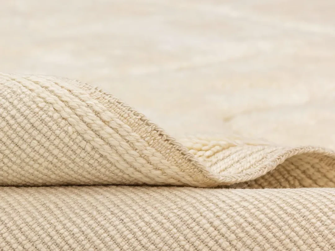 Close-up of the edge of a beige woven rug, showing its textured surface and slightly lifted corner.