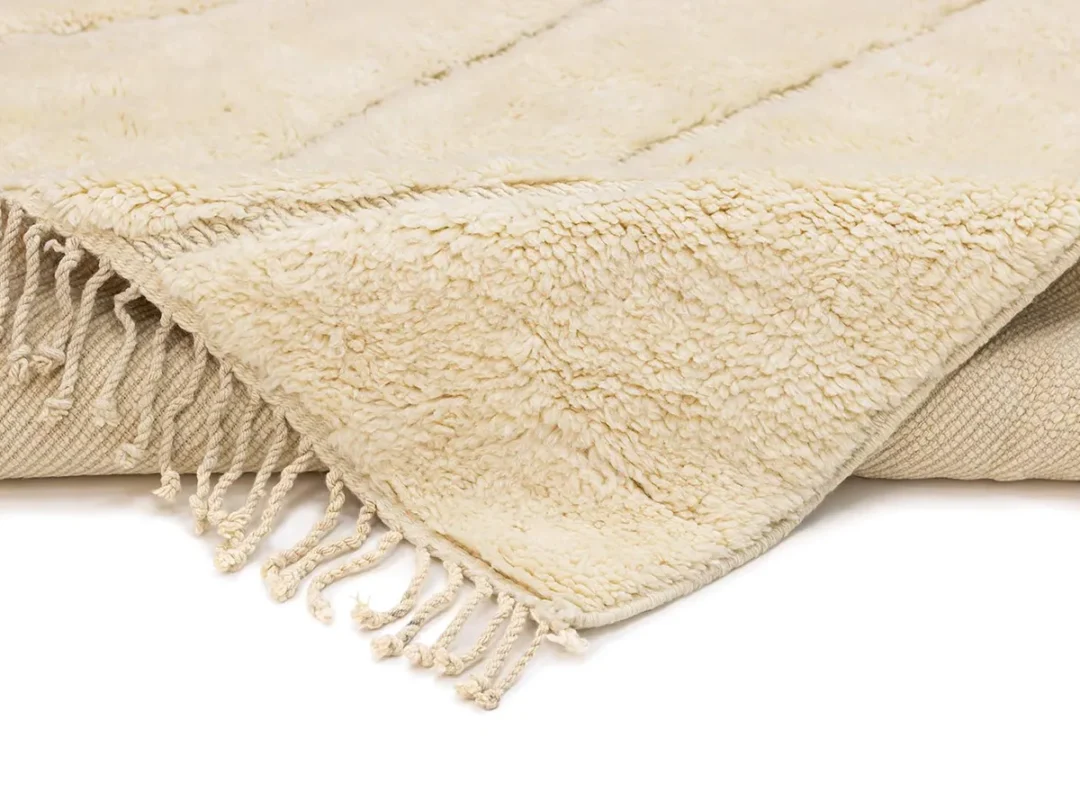 A close-up of a beige, textured rug with fringed edges, partially folded over to show the underside.