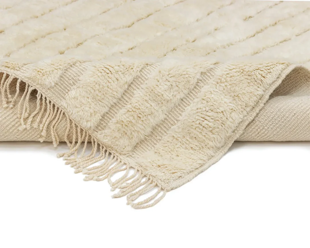 A close-up view of a cream-colored, textured rug with fringe along the edges, partially folded over on a white background.