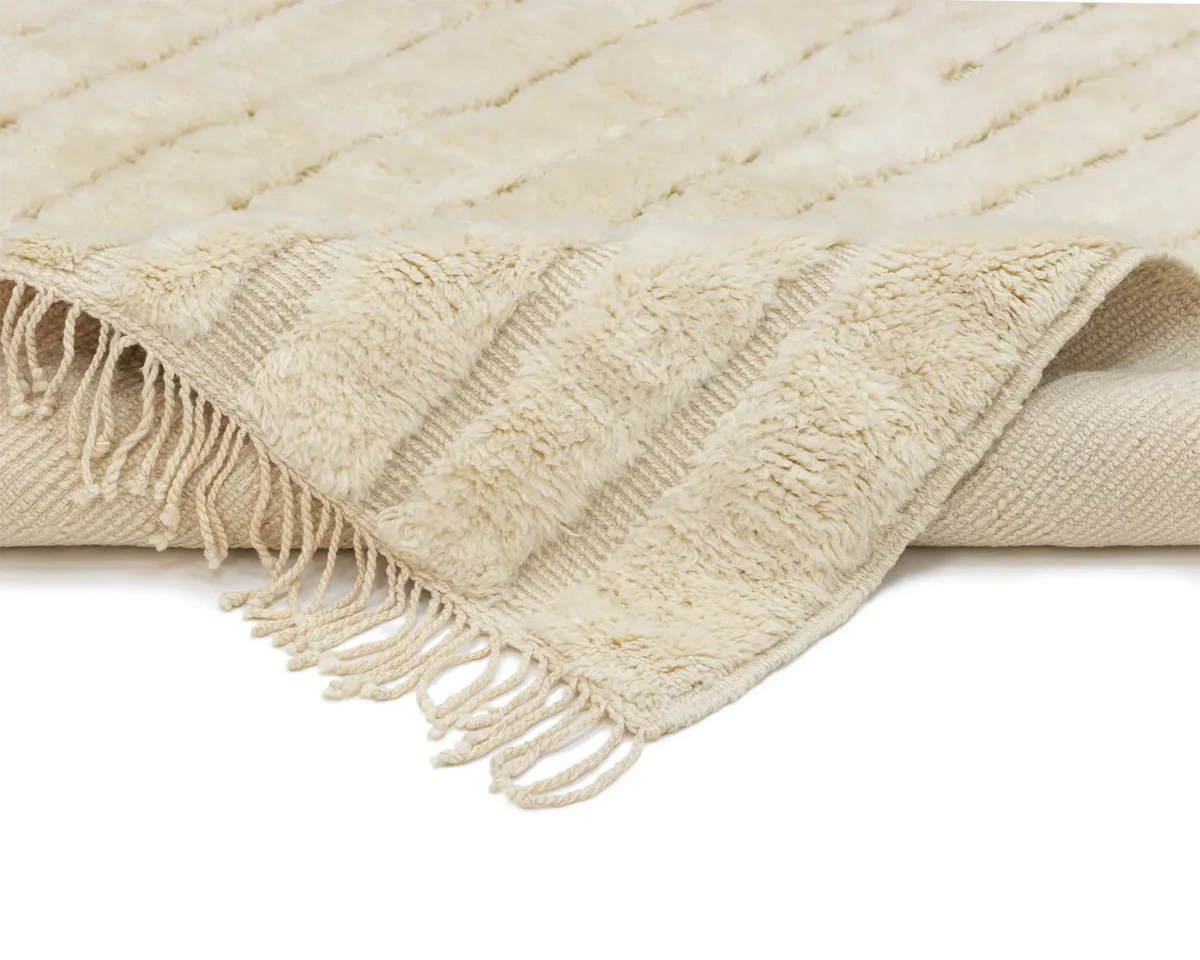 A close-up view of a cream-colored, textured rug with fringe along the edges, partially folded over on a white background.