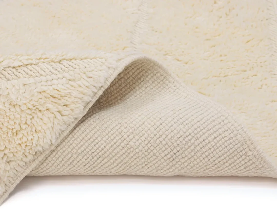 Close-up of a cream-colored, textured rug with one corner folded to show the woven backing and soft, fluffy pile on top.