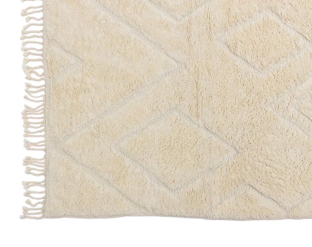 A section of a cream-colored rug with a subtle diamond pattern and fringe on the left edge.