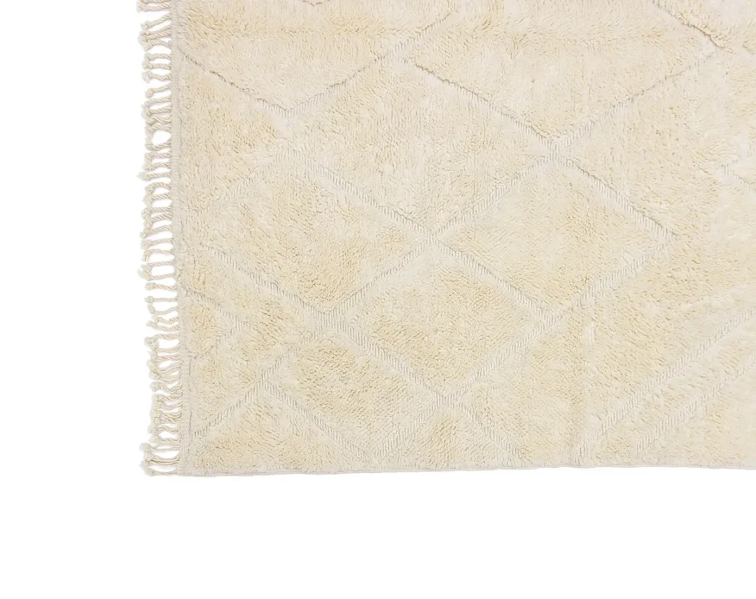 A close-up view of a beige textured rug with a diamond pattern and fringe along the left edge.