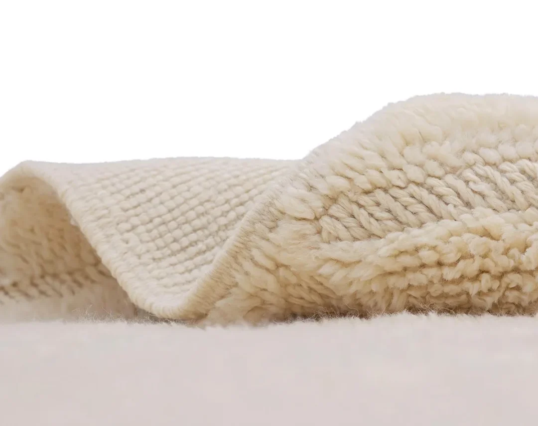 Close-up of a cream-colored, textured, and folded wool rug against a plain white background.