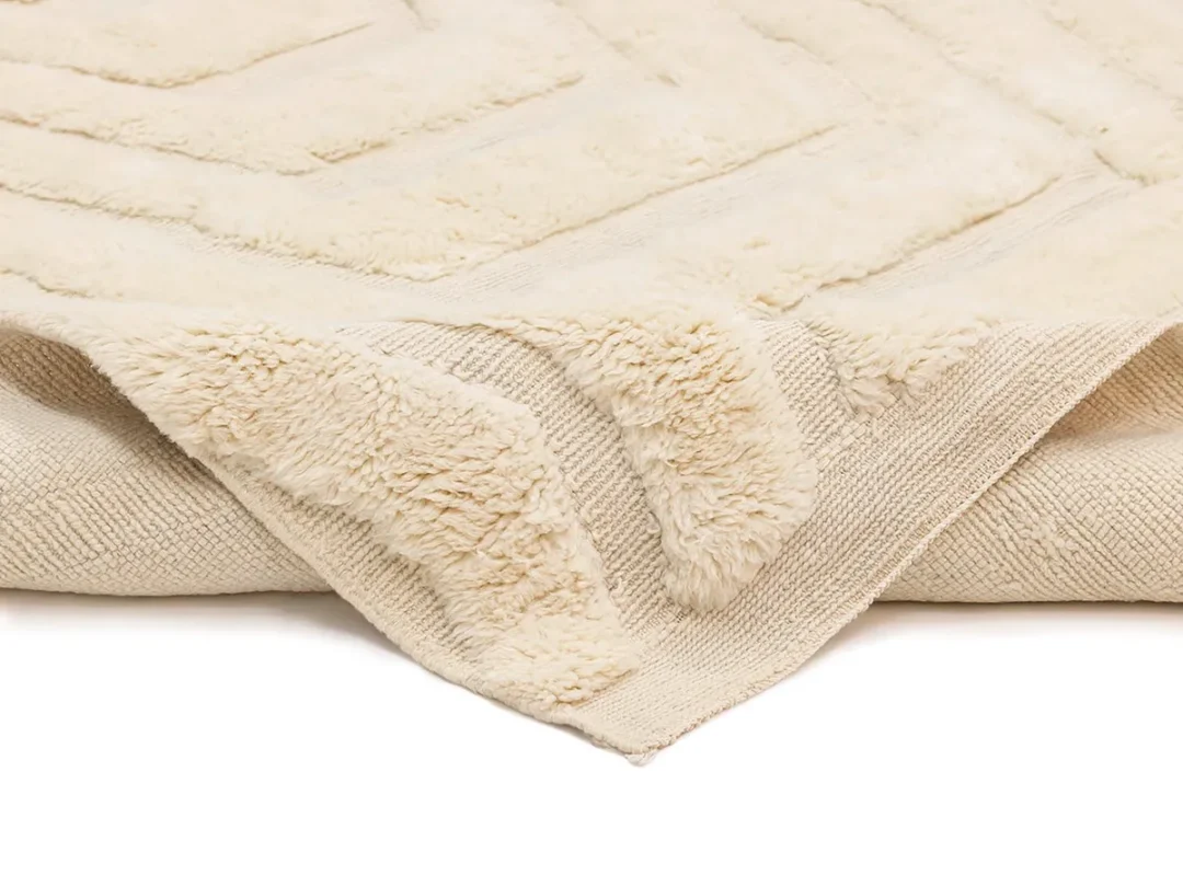 A close-up view of a beige, textured rug with a soft, plush surface and a patterned border, laid flat on a white background.
