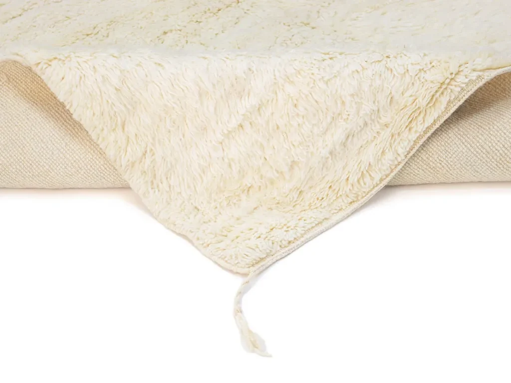 A close-up of a cream-colored, textured rug with a corner folded over, showing soft, fuzzy material and a loose strand.