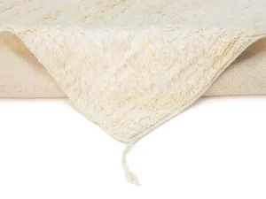 A close-up of a cream-colored, textured blanket or rug with a corner folded over, showing soft, fuzzy material and a loose strand.