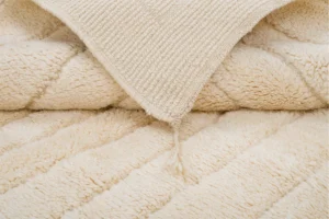 Close-up of a folded cream-colored textured blanket with a diagonal ribbed pattern and tassel at the corner.