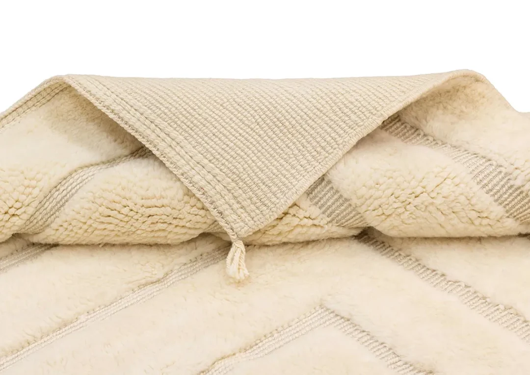 Folded cream-colored textured rug with a tassel on the corner, featuring a geometric pattern and a visible woven underside.