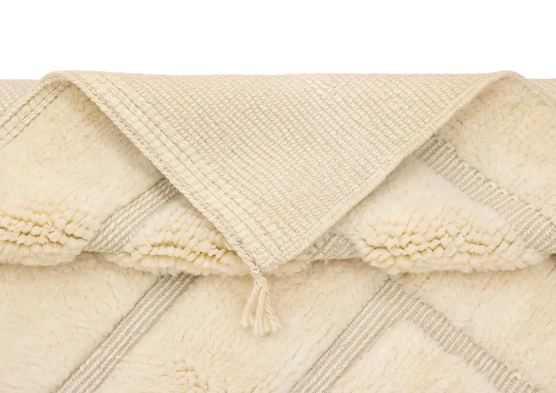 A close-up of a folded cream-colored textured rug with diagonal lines and a tassel at the edge, set against a white background.