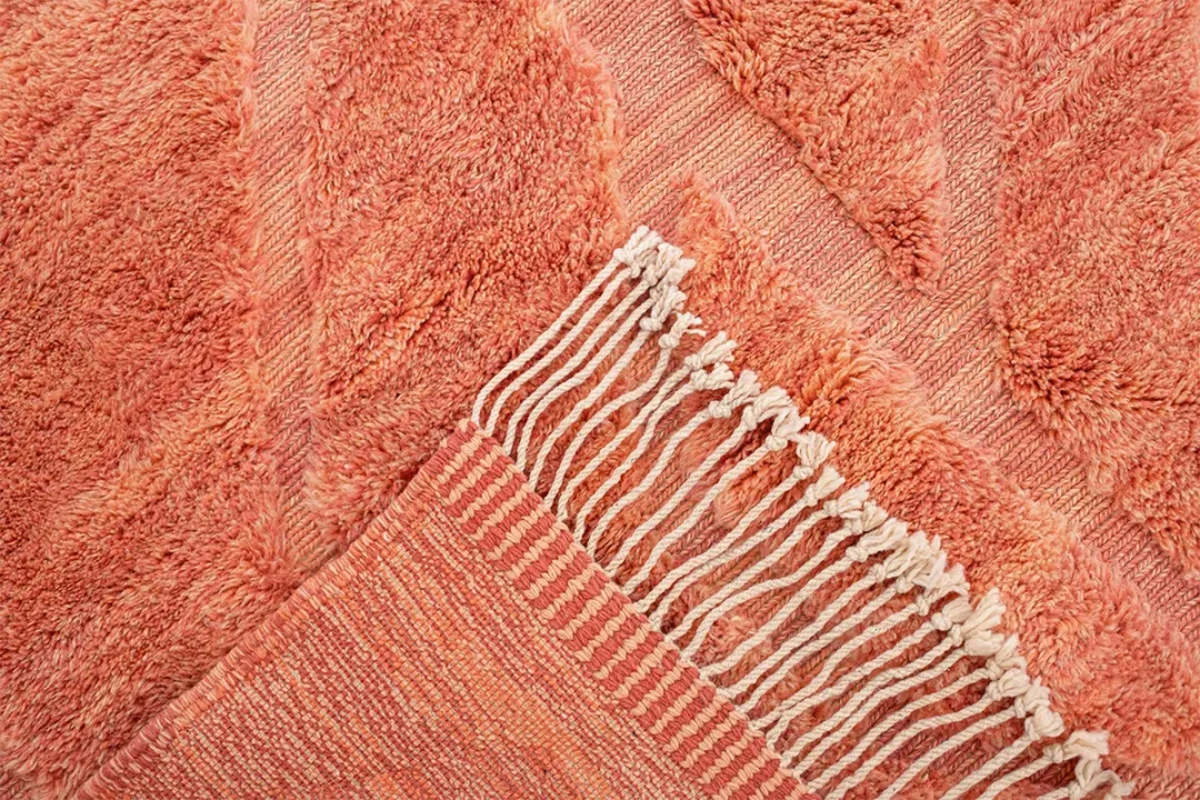 Close-up of a textured, coral-colored rug with geometric patterns and white fringes along one edge.