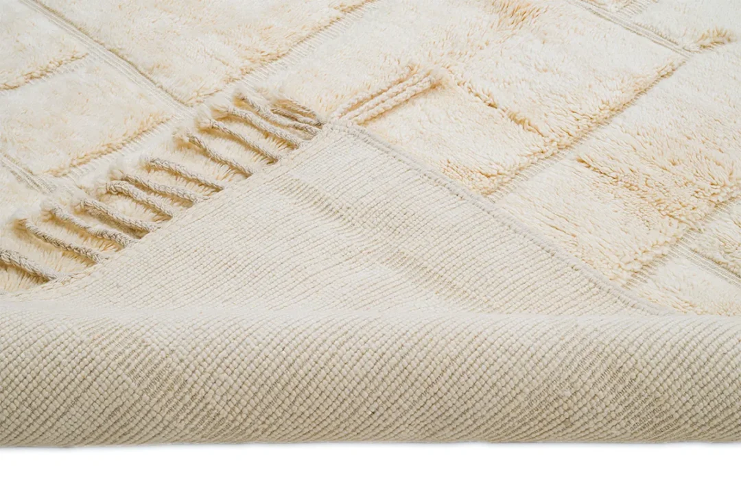 A close-up of a beige textured rug with a checkered pattern, showing one corner folded over to reveal the woven underside and tassel fringe.