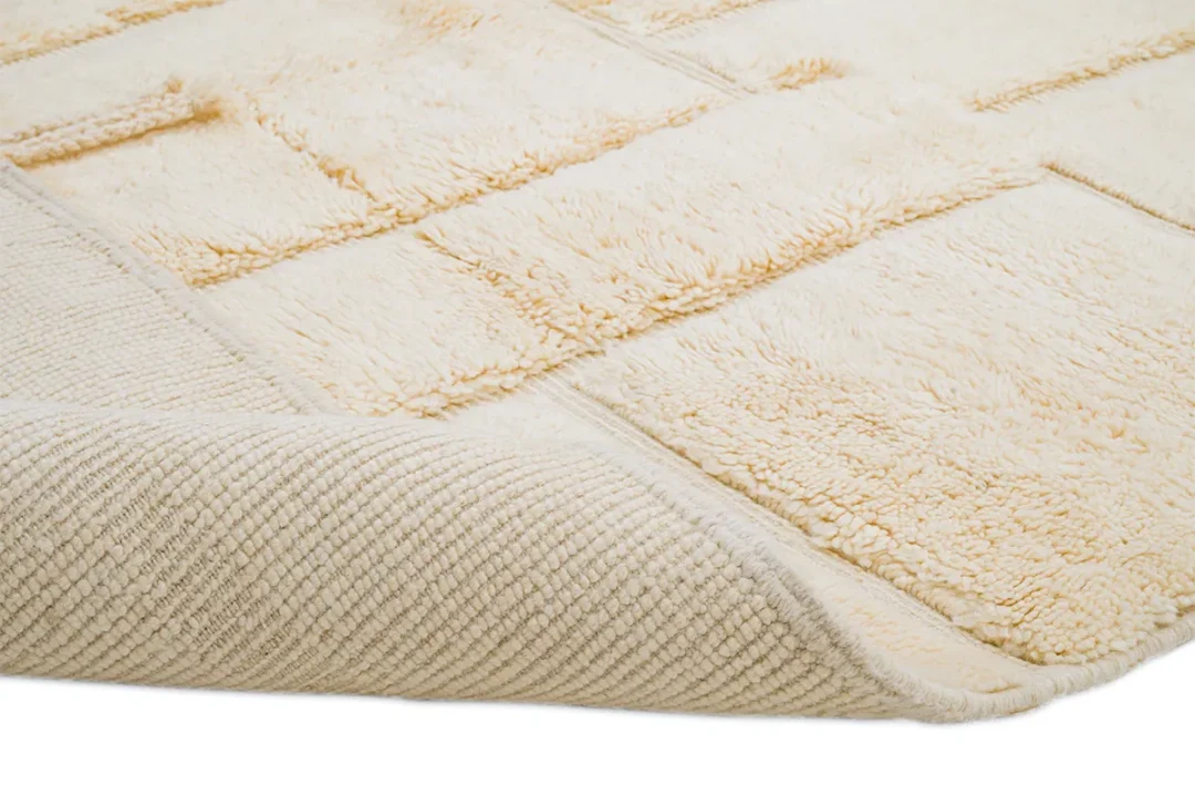 Close-up of a beige textured rug with a raised geometric pattern and one corner slightly folded to show the underside.