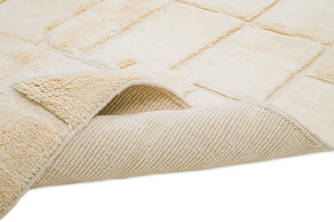 A close-up of a beige textured rug with one corner slightly folded to reveal the woven backing underneath.