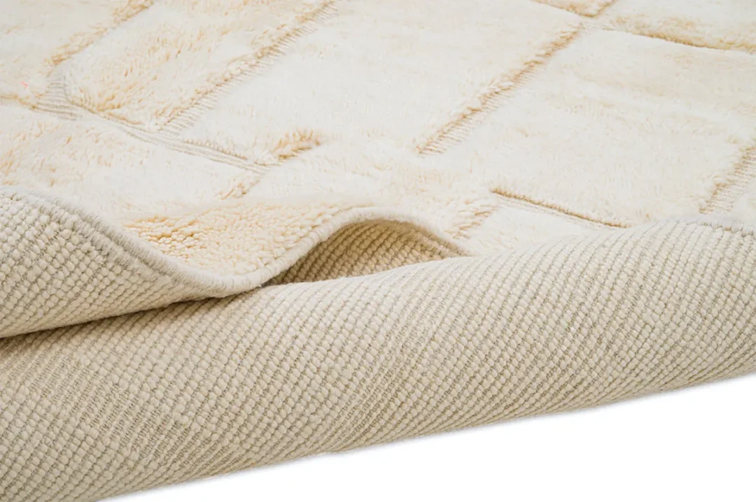 A close-up of a beige textured rug with a soft, plush surface and a woven underside, slightly folded at one corner.