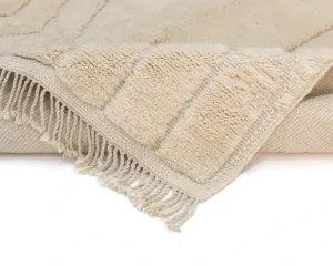 A close-up of a beige textured rug with fringe detailing on the edge, showing a soft and plush surface.
