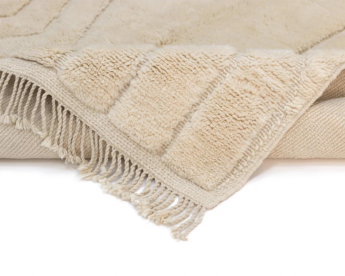 A close-up of a beige textured rug with fringe detailing on the edge, showing a soft and plush surface.
