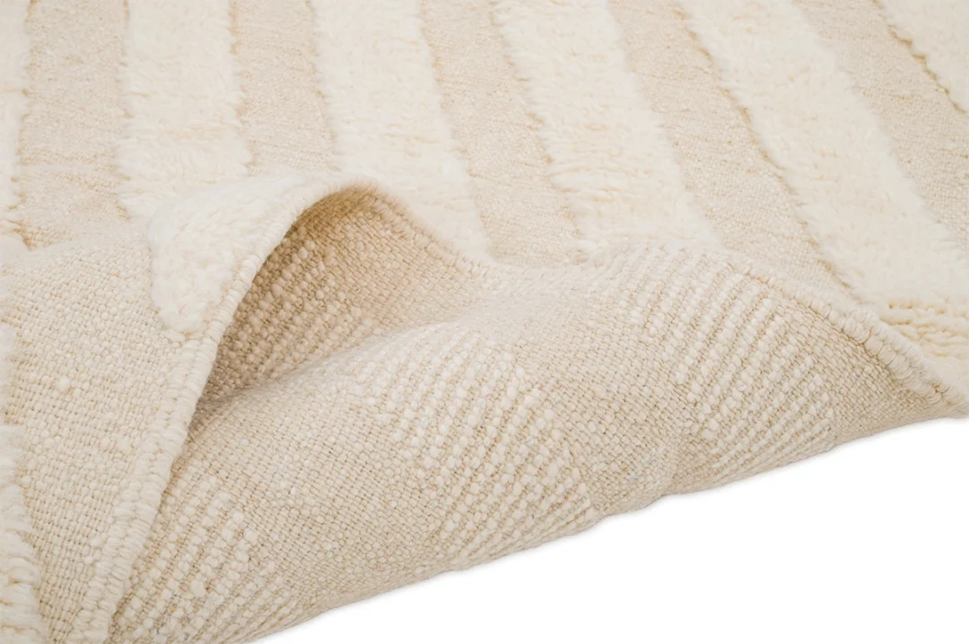 A close-up view of a cream-colored textured rug with raised, striped patterns, showing one corner slightly folded over.