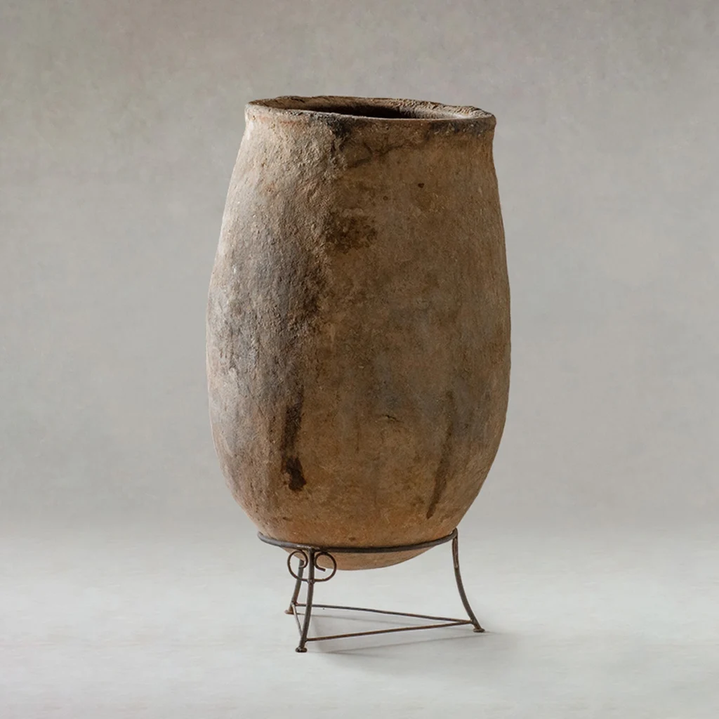 Large, weathered clay vessel with a rough surface, positioned upright on a simple metal stand against a neutral background.