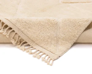 Close-up of a cream-colored, textured blanket with a soft, plush surface and fringed edges, partially folded on a flat surface.